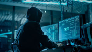 Shot from the Back to Hooded Hacker Breaking into Corporate Data Servers from His Underground Hideout. Place Has Dark Atmosphere, Multiple Displays, Cables Everywhere.